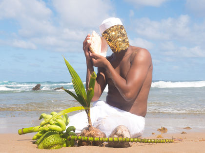 A Black non-binary person sits on a sandy beach in front of the sea and a blue and white clouded sky. They are wearing a rimless white hat and white fabric covers their legs and their chest is bare. Their face is covered by a gold sequin veil. They are delicately holding a shell to one ear and there is an array of Caribbean fruits around them.
