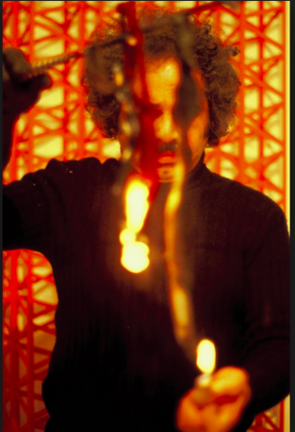 Rasheed, a Pakistani man, is pictured in a black jumper holding a baton from which two tails are hanging down and covering his eyes. The left tail is on fire and he holds a flame to the bottom of the other tail. The background uses similar colours to the fire, with red, scaffolding-like beams criss-crossing over a yellow background.